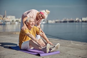 high-spirited-pensioner-performing-seated-forward-bend-on-the-mat-assisted-by-his-cheerful-wife
